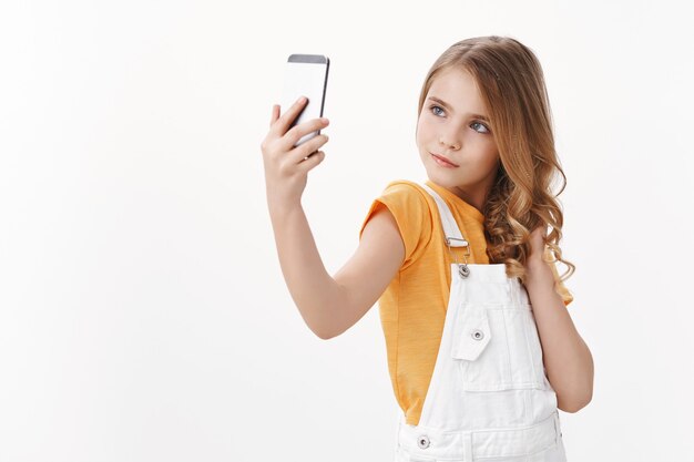 Sassy glamour linda niña bonita con cabello rubio sostenga el teléfono inteligente, tomando selfie posando femenina y tonta, haciendo pucheros mirada confiada, imitando a mujeres adultas, de pie en la pared blanca