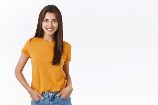 Sassy guapa mujer morena femenina en camiseta amarilla se toma de la mano en los bolsillos de los jeans sonriendo segura de sí misma con una sonrisa blanca perfecta de pie fondo blanco carismática y entusiasta