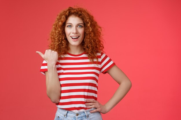 Sassy guapo emotivo feliz sonriente pelirroja europea peinado rizado femenino apuntando con el pulgar hacia la izquierda sonrisa asertiva descarada mantenga la cintura de la mano dirigiendo la oferta de publicidad promocional fondo rojo.