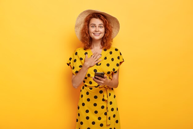 Satisfecha e impresionada mujer pelirroja toca el pecho, usa sombrero de verano y vestido de lunares amarillos