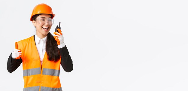 Satisfecha, feliz y sonriente ingeniera asiática, técnica industrial con casco de seguridad y uniforme que muestra el pulgar hacia arriba mientras elogia el gran trabajo usando walkietalkie, da permiso para trabajar