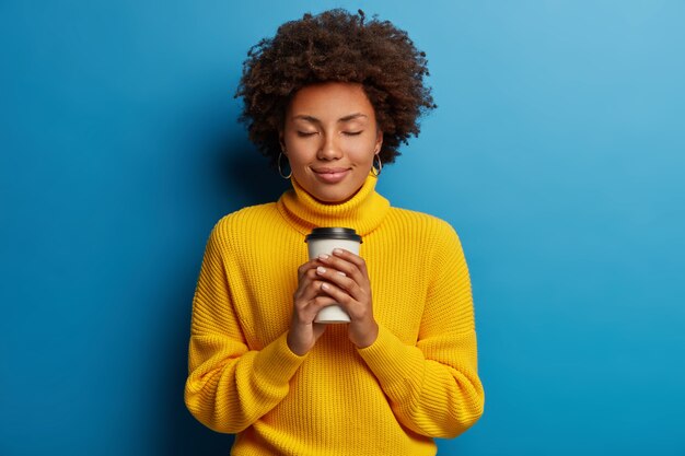 Satisfecha joven mujer afro adora la bebida, sostiene café para llevar, cierra los ojos, viste un suéter amarillo, aislado sobre fondo azul.