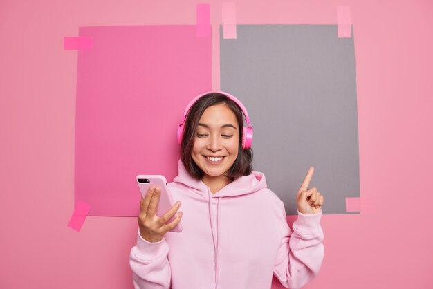 Satisfecha mujer asiática milenaria hace videollamadas promociona algo indica en el espacio en blanco sonríe agradablemente muestra dirección venta logo banner de la tienda usa poses de sudadera con capucha contra la pared rosa