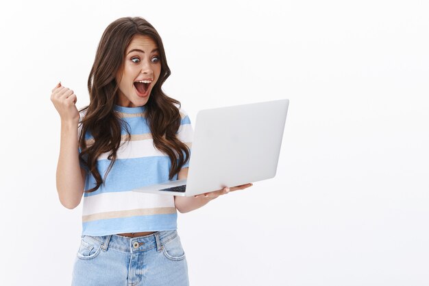 Satisfecha mujer moderna feliz y afortunada recibir noticias excelentes a través del buzón de correo, bomba de puño, sostener la computadora portátil divertida, leer un mensaje de texto impresionante, mirar la pantalla de la computadora, terminar el trabajo triunfante, pared blanca