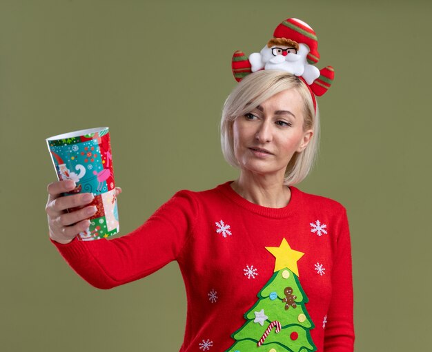 Satisfecha mujer rubia de mediana edad con diadema de santa claus y suéter navideño extendiendo la taza plástica de navidad hacia la cámara mirándolo aislado en la pared verde oliva