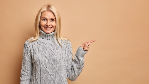 Satisfecha rubia mujer de mediana edad con arrugas viste suéter gris cálido, apuntando al espacio de copia