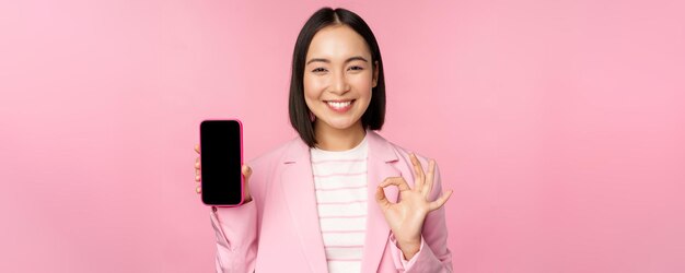 Satisfecha y sonriente mujer de negocios asiática recomendando la compañía del sitio web de la aplicación de teléfono móvil en el teléfono inteligente que muestra la pantalla y el fondo rosa de la señal OK