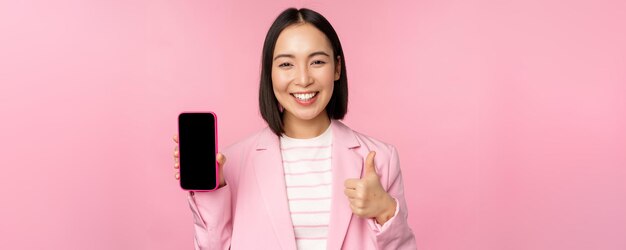 Satisfecha y sonriente mujer de negocios asiática recomendando la empresa del sitio web de la aplicación de teléfono móvil en el teléfono inteligente que muestra la pantalla y el pulgar hacia arriba con fondo rosa