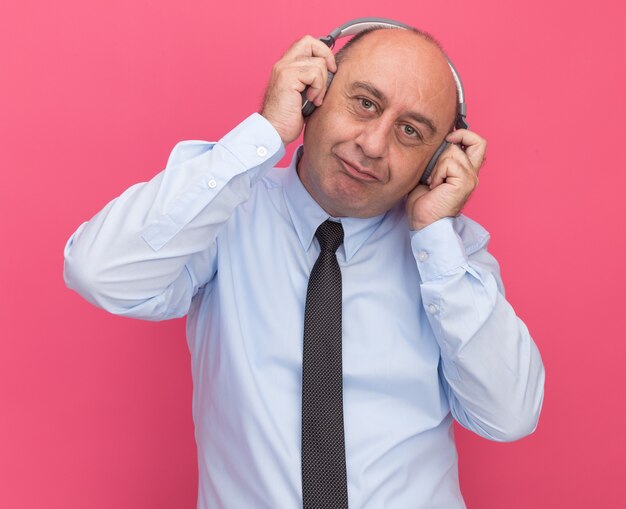 Satisfecho con la cabeza inclinada hombre de mediana edad vestido con camiseta blanca con corbata y auriculares aislados en la pared rosa