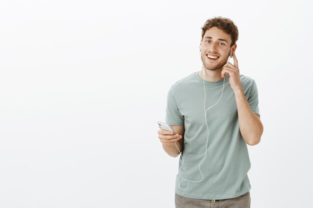 Satisfecho complacido modelo masculino caucásico con cerdas, sosteniendo el teléfono inteligente y tocando el auricular mientras escucha música y disfruta del agradable sonido de los auriculares