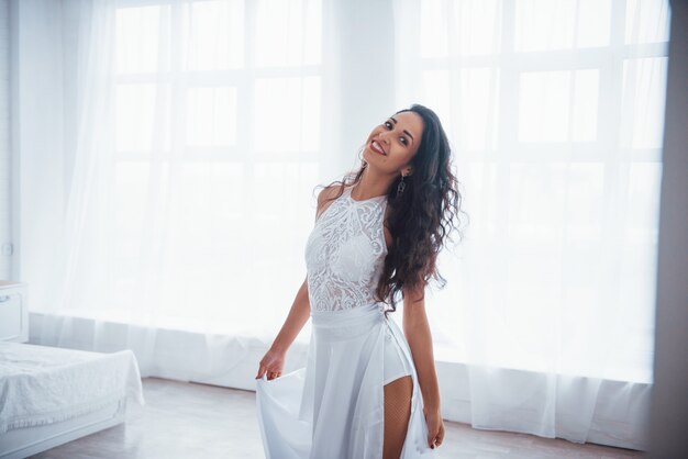 Foto gratuita satisfecho y gratis. hermosa mujer vestida de blanco se encuentra en la sala blanca con luz del día a través de las ventanas