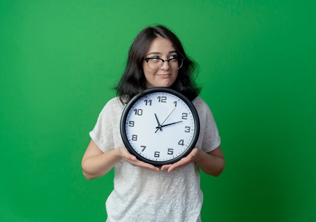 Satisfecho joven bastante caucásica con gafas sosteniendo reloj mirando al lado aislado sobre fondo verde con espacio de copia