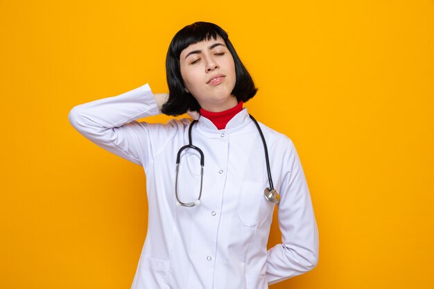 Satisfecho joven bastante caucásica en uniforme médico con estetoscopio poniendo la mano sobre su cabeza de pie con los ojos cerrados