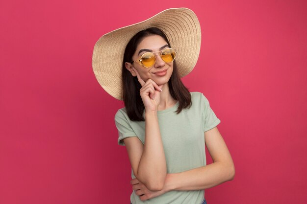 Satisfecho joven bastante caucásica vestida con sombrero de playa y gafas de sol poniendo la mano en el mentón aislado en la pared rosa con espacio de copia