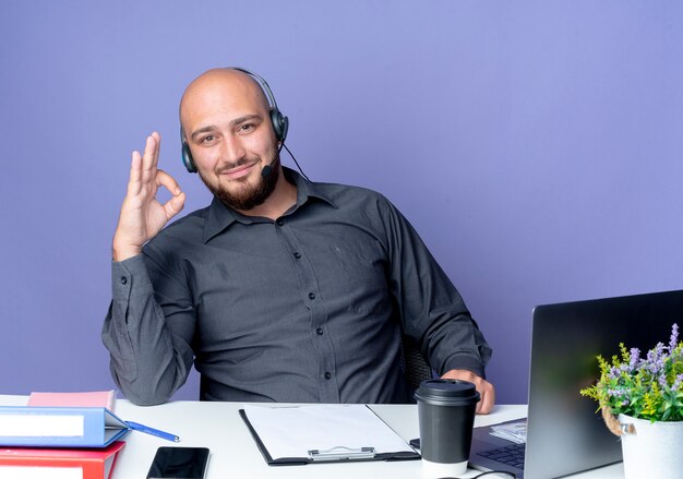 Satisfecho joven calvo call center hombre con auriculares sentado en un escritorio con herramientas de trabajo haciendo bien firmar aislado sobre fondo púrpura