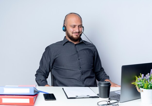 Satisfecho joven calvo call center hombre con auriculares sentado en un escritorio con herramientas de trabajo mirando portátil aislado sobre fondo blanco.