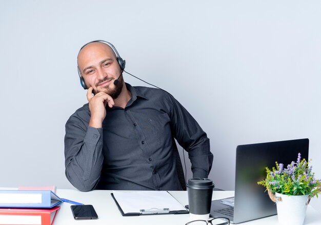 Satisfecho joven calvo call center hombre con auriculares sentado en el escritorio con herramientas de trabajo poniendo el dedo en la mejilla aislado sobre fondo blanco.