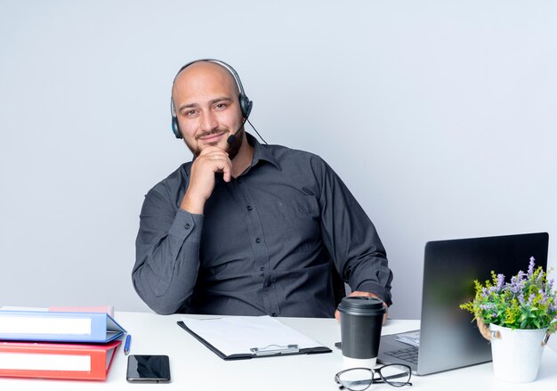 Satisfecho joven calvo call center hombre con auriculares sentado en el escritorio con herramientas de trabajo poniendo la mano en el mentón aislado sobre fondo blanco.