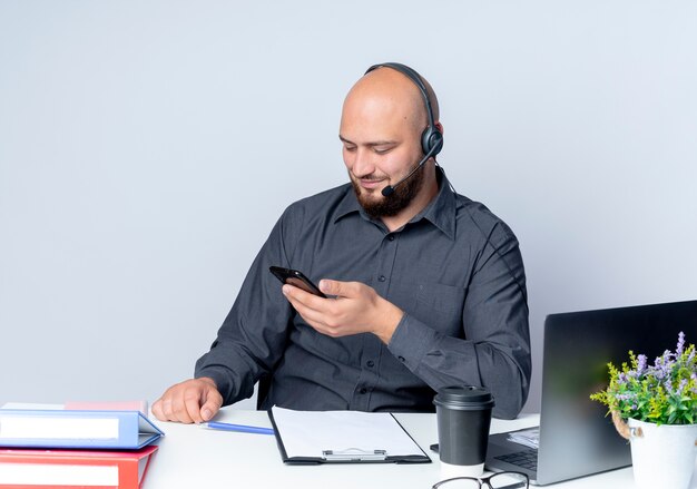 Satisfecho joven calvo call center hombre con auriculares sentado en el escritorio con herramientas de trabajo sosteniendo y mirando el teléfono móvil aislado sobre fondo blanco.