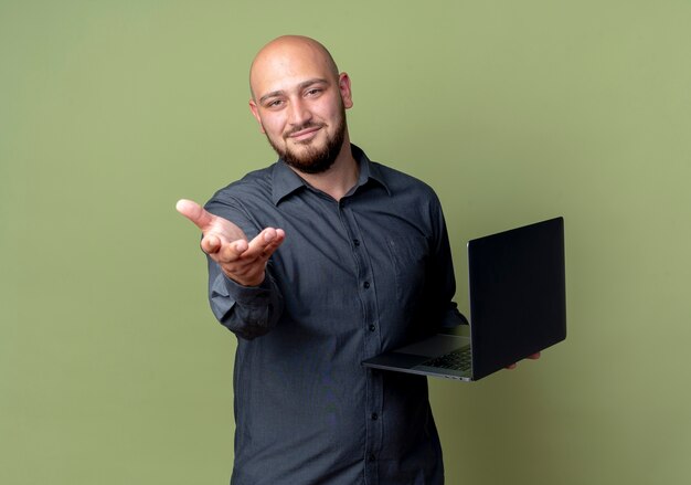 Satisfecho joven calvo call center hombre sujetando un portátil y estirando la mano a la cámara aislada sobre fondo verde oliva con espacio de copia