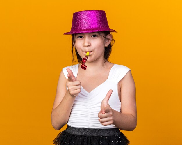 Satisfecho joven caucásica con gorro de fiesta púrpura soplando un silbato de fiesta y apuntando aislado en la pared naranja con espacio de copia