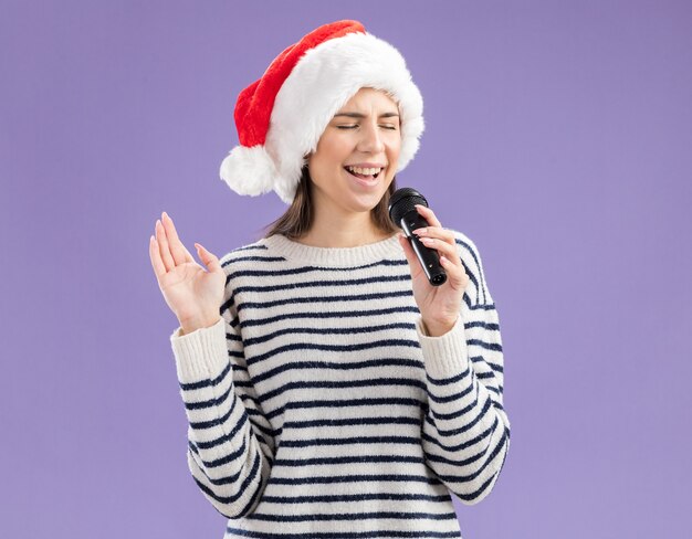 Satisfecho joven caucásica con gorro de Papá Noel tiene micrófono fingiendo cantar aislado sobre fondo púrpura con espacio de copia