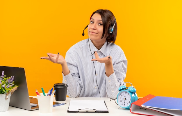 Satisfecho joven centro de llamadas con auriculares sentado en el escritorio mostrando las manos vacías aisladas sobre fondo naranja