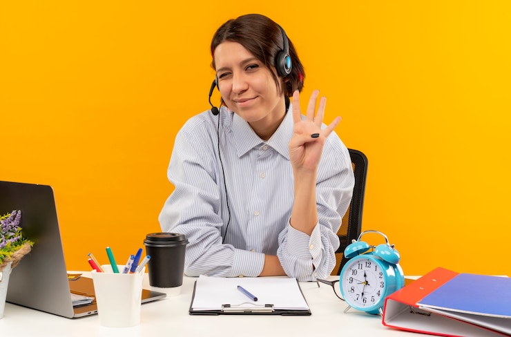 Foto gratuito satisfecho joven chica del centro de llamadas con auriculares sentado en el escritorio guiñando un ojo y mostrando cuatro con la mano aislada sobre fondo naranja