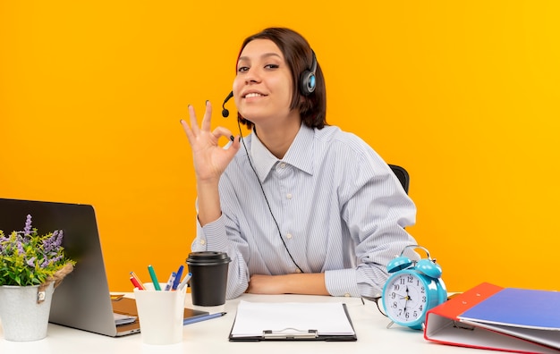 Satisfecho joven chica del centro de llamadas con auriculares sentados en el escritorio haciendo bien firmar aislado sobre fondo naranja