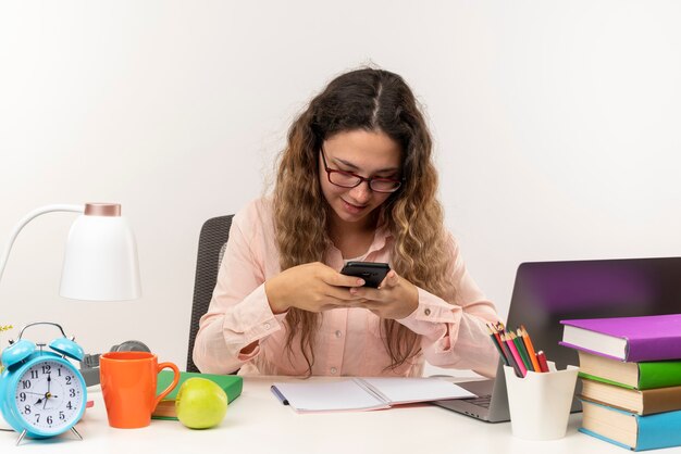 Satisfecho joven colegiala bonita con gafas sentado en el escritorio con herramientas escolares haciendo sus deberes usando su teléfono aislado en la pared blanca