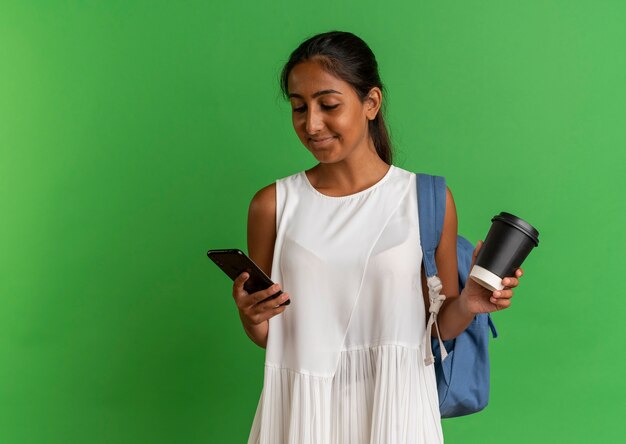 Satisfecho joven colegiala vistiendo bolsa trasera sosteniendo una taza de café y mirando el teléfono en la mano