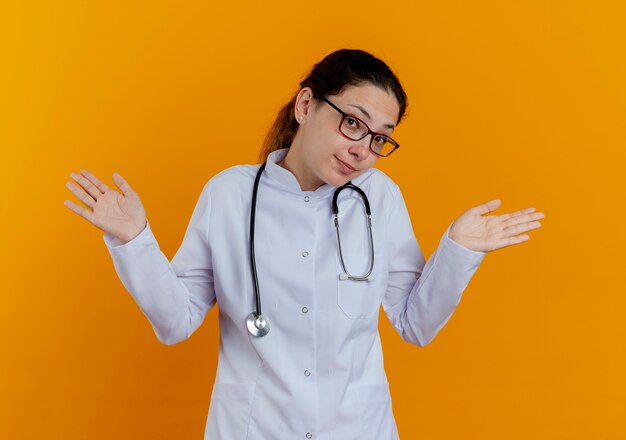 Satisfecho joven doctora vistiendo bata médica y estetoscopio con gafas extendiendo las manos aisladas en la pared naranja