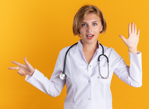 Satisfecho joven doctora vistiendo bata médica con estetoscopio mostrando diferentes números aislados en la pared naranja