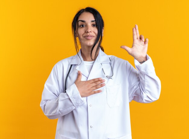Satisfecho joven doctora vistiendo bata médica con estetoscopio mostrando gesto de pistola aislado en la pared amarilla