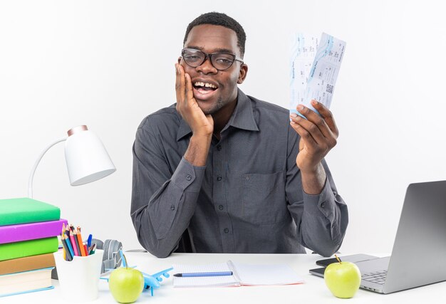 Satisfecho joven estudiante afroamericano con gafas ópticas sentado en el escritorio con herramientas escolares poniendo la mano en la cara y sosteniendo boletos de avión aislados en la pared blanca