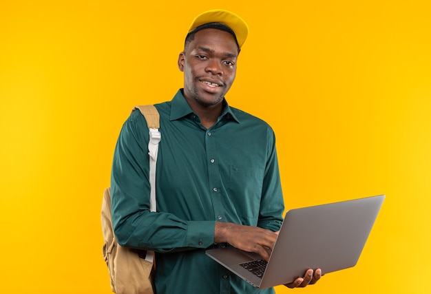 Satisfecho joven estudiante afroamericano con gorra y mochila sosteniendo portátil