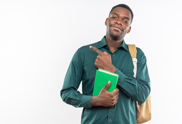 Satisfecho joven estudiante afroamericano con mochila sosteniendo libro y apuntando a un lado