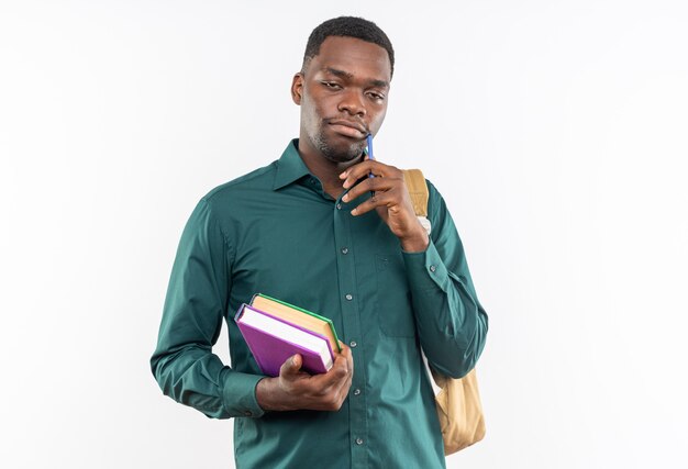 Satisfecho joven estudiante afroamericano con mochila sosteniendo libros y bolígrafo