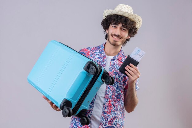 Satisfecho joven guapo viajero rizado hombre con sombrero sosteniendo billetera y boletos de avión y maleta en espacio en blanco aislado con espacio de copia