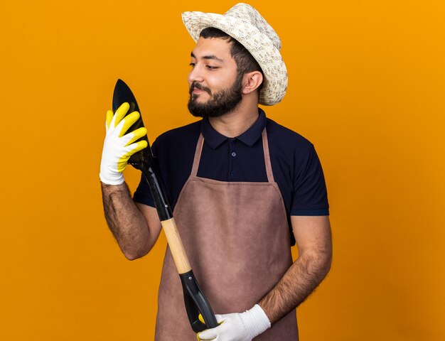 Satisfecho joven jardinero varón caucásico vistiendo gorro y guantes de jardinería sosteniendo y mirando la pala aislado en la pared naranja con espacio de copia