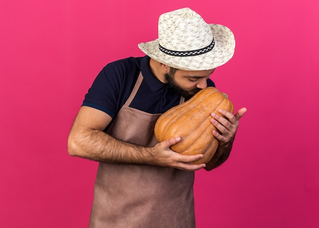 Satisfecho joven jardinero varón caucásico vistiendo sombrero de jardinería sosteniendo y besando calabaza aislado en la pared rosa con espacio de copia