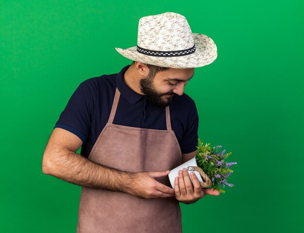 Satisfecho joven jardinero varón caucásico vistiendo sombrero de jardinería sosteniendo y mirando maceta aislado en la pared verde con espacio de copia