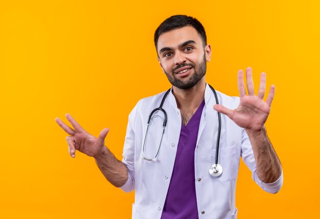 Satisfecho joven médico con estetoscopio bata médica mostrando diferentes números con las manos sobre fondo amarillo aislado