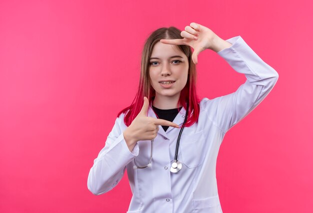 Satisfecho joven médico con estetoscopio bata médica mostrando gesto de marco sobre fondo rosa aislado