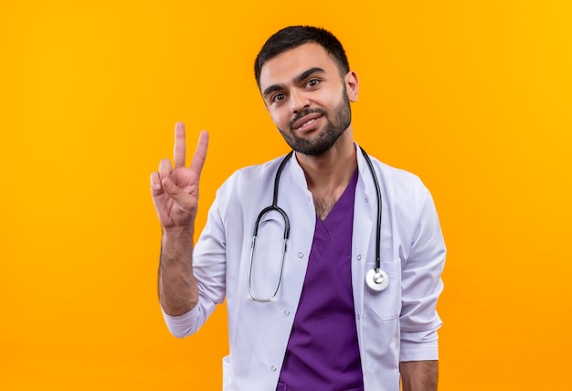 Satisfecho joven médico con estetoscopio bata médica mostrando gesto de paz sobre fondo amarillo aislado