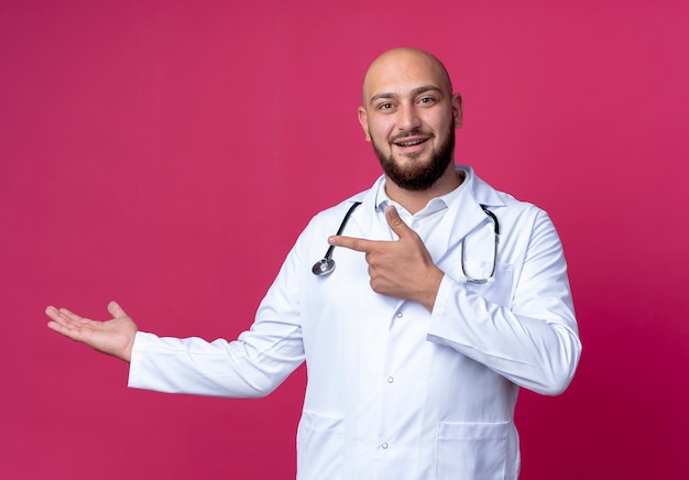 Satisfecho joven médico varón calvo vistiendo bata médica y estetoscopio fingiendo sostener algo aislado sobre fondo rosa con espacio de copia