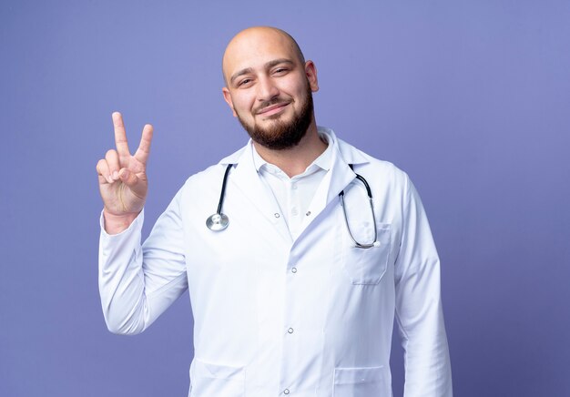 Satisfecho joven médico varón calvo vistiendo bata médica y estetoscopio mostrando gesto okey aislado sobre fondo azul.