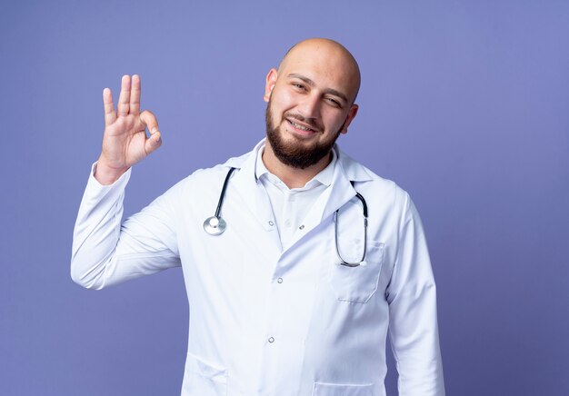 Satisfecho joven médico varón calvo vistiendo bata médica y estetoscopio mostrando gesto okey aislado sobre fondo azul.