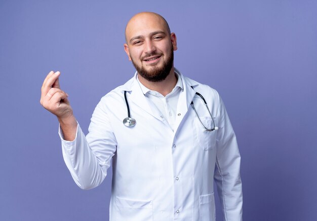 Satisfecho joven médico varón calvo vistiendo bata médica y estetoscopio mostrando gesto de punta aislado sobre fondo azul.