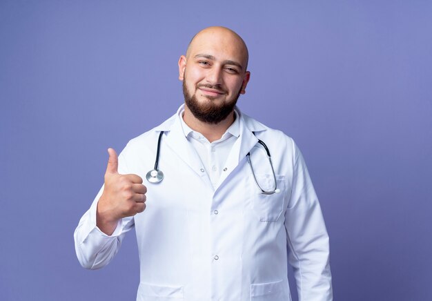 Foto gratuita satisfecho joven médico varón calvo vistiendo bata médica y estetoscopio con el pulgar hacia arriba aislado sobre fondo azul.